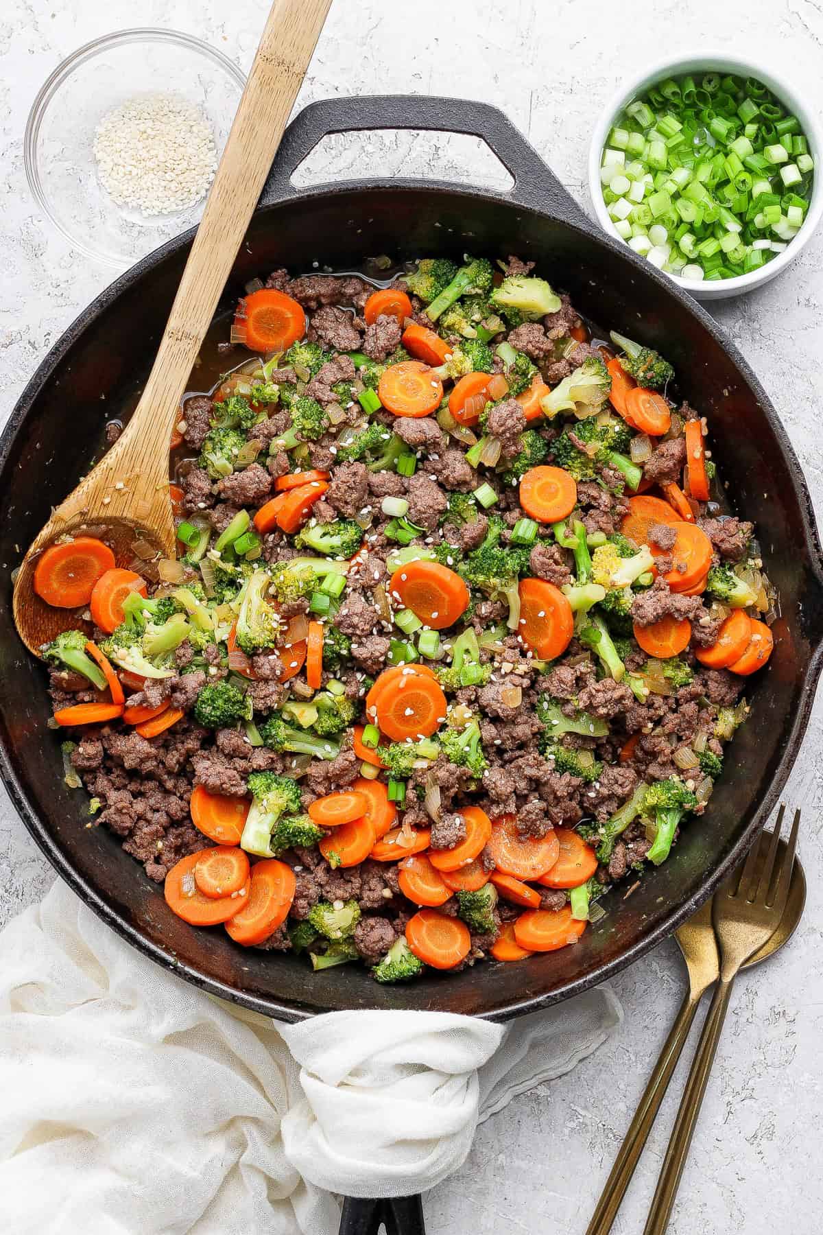 A skillet filled with cooked ground beef, sliced carrots, broccoli, and green onions. A wooden spoon rests inside the skillet, with bowls of sesame seeds and chopped green onions nearby.