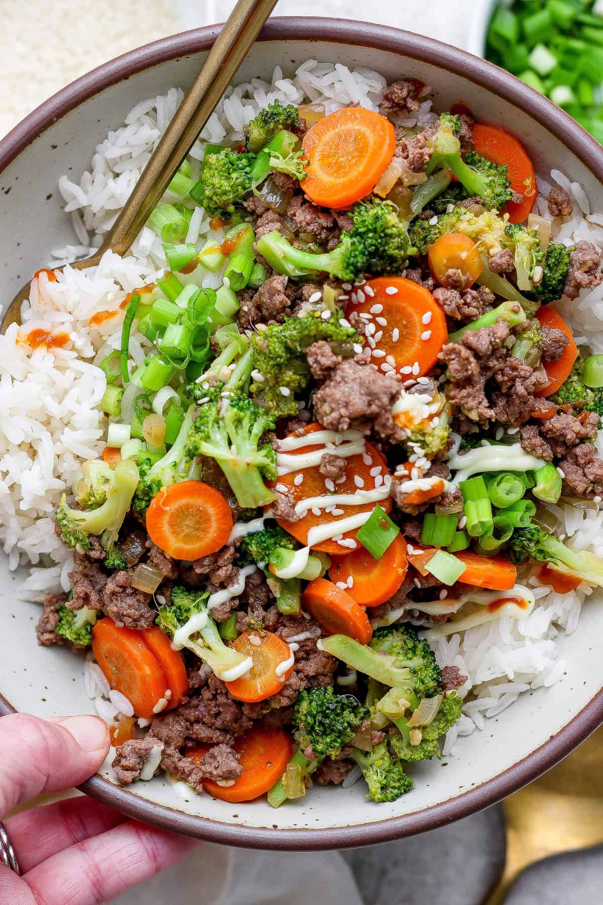A bowl of white rice topped with ground beef, sliced carrots, broccoli, green onions, and sesame seeds, with chopsticks resting on the edge.