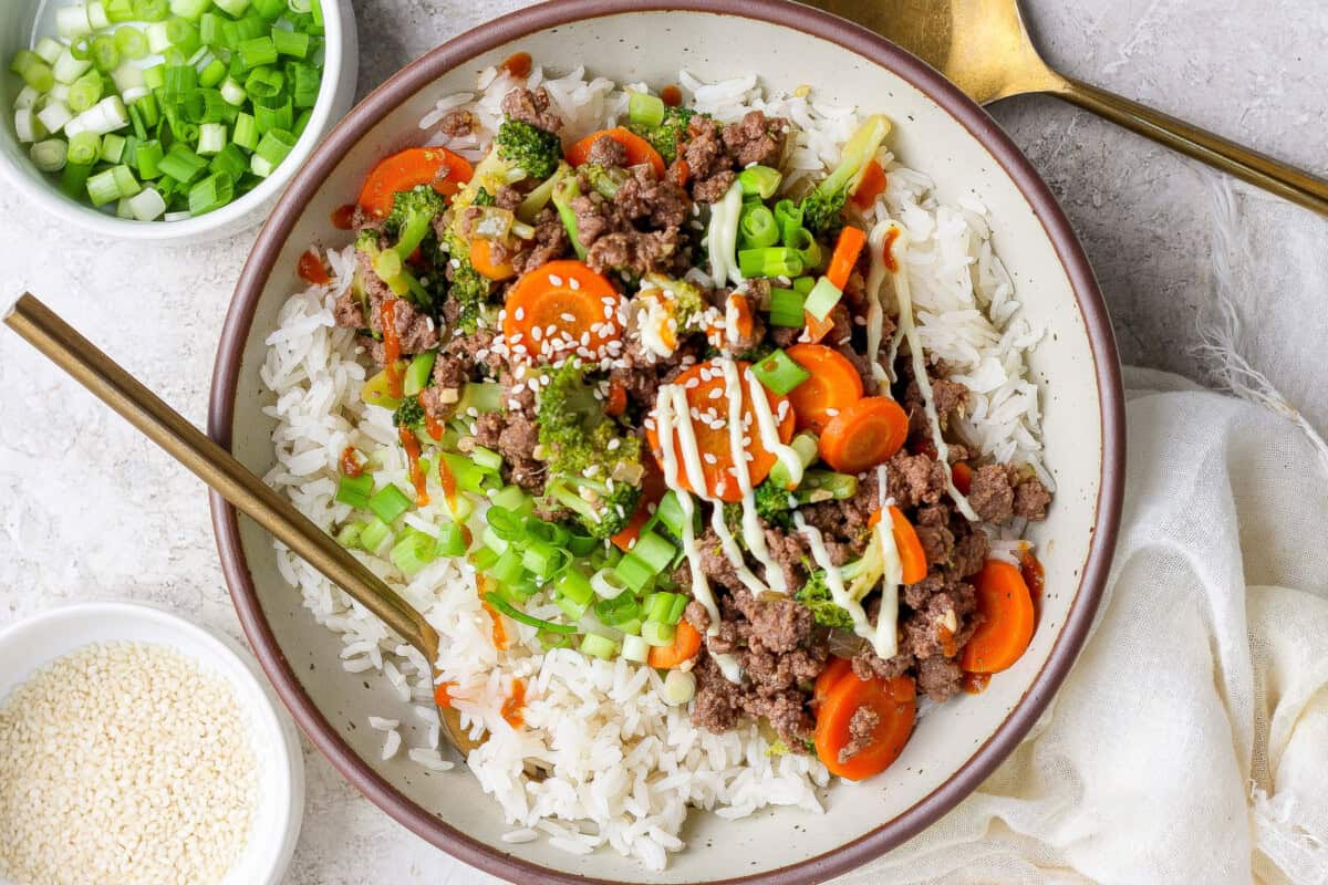 A bowl of white rice topped with ground beef, sliced carrots, broccoli, chopped green onions, sesame seeds, and a drizzle of sauce. Spoons and small bowls of green onions and sesame seeds nearby.