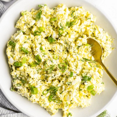 A bowl of egg salad with chopped green onions and dill, accompanied by a gold spoon.