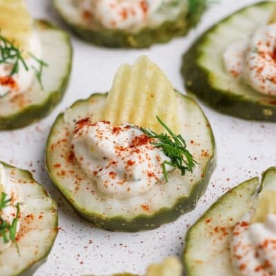 Cucumber slices topped with sour cream, a potato chip, dill, and paprika on a white surface.