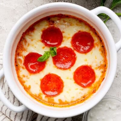 Baked dish with melted cheese and six pepperoni slices, garnished with basil, in a white ceramic bowl on a textured surface.