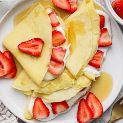 Crepes folded and topped with sliced strawberries, cream, and syrup on a white plate.