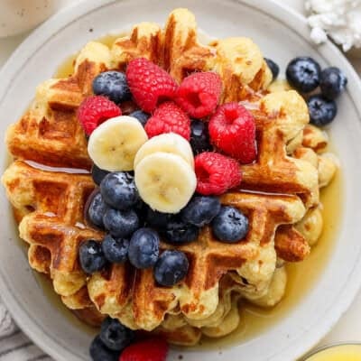 A stack of waffles topped with blueberries, raspberries, banana slices, and drizzled with syrup on a round white plate.