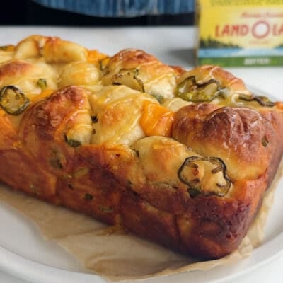 A loaf of pull-apart bread with melted cheese and sliced jalape&ntilde;os on parchment paper, with a box of butter in the background.