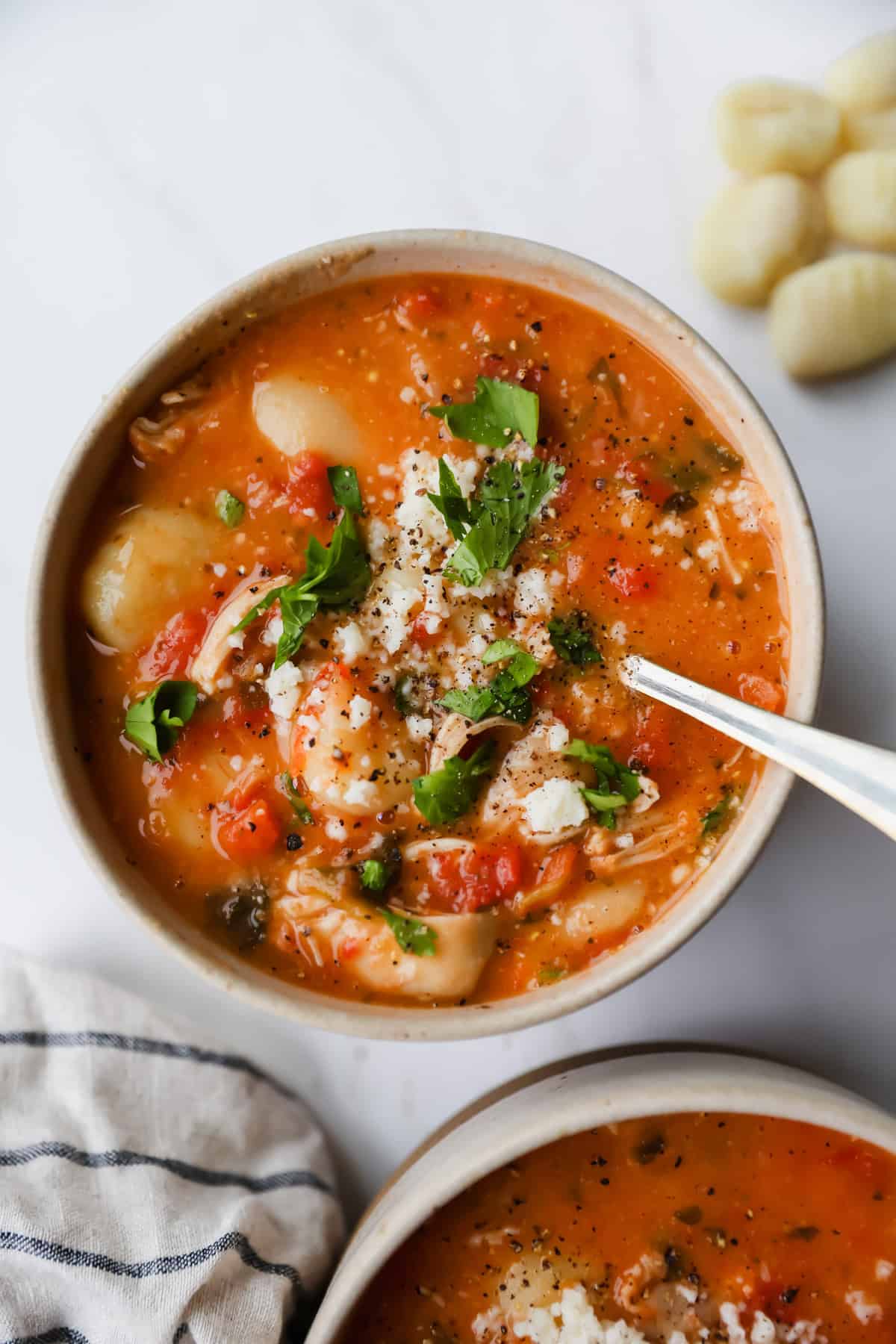 A bowl of tomato-based soup with gnocchi, shredded chicken, herbs, and cheese, with a spoon in the bowl and several gnocchi pieces and a napkin nearby.