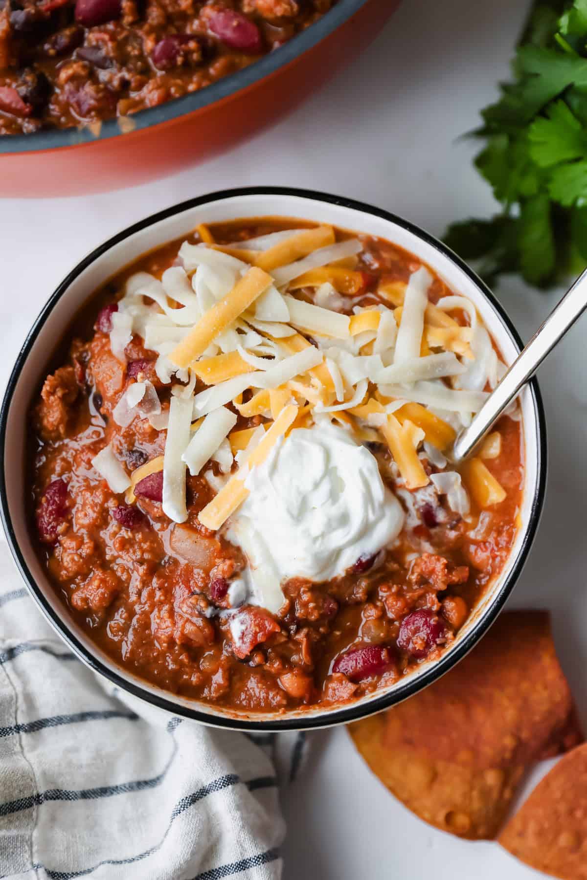 A bowl of chili topped with shredded cheese and a dollop of sour cream, with a spoon, tortilla chips, and a striped napkin beside it.
