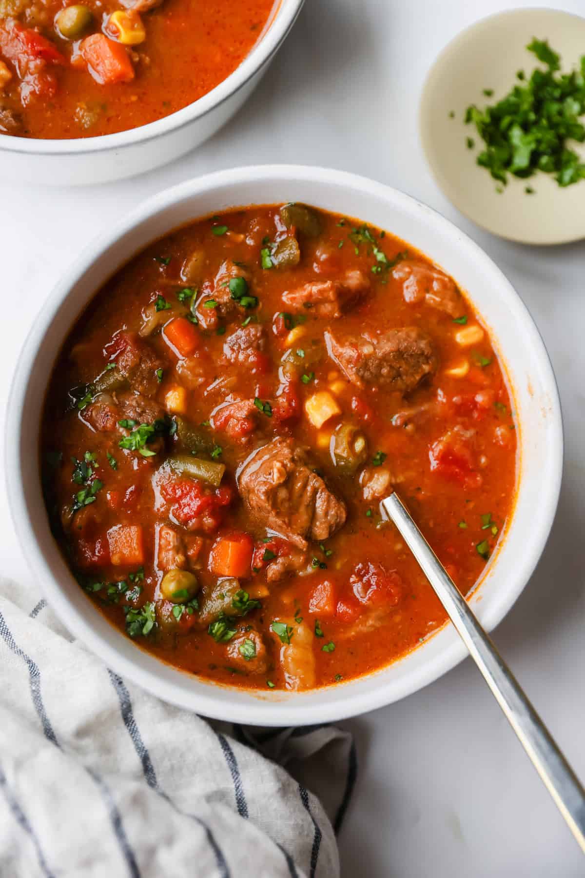 A bowl of beef and vegetable soup with a spoon, featuring chunks of meat, peas, carrots, corn, and tomatoes. A plate of chopped herbs sits nearby on a white surface.