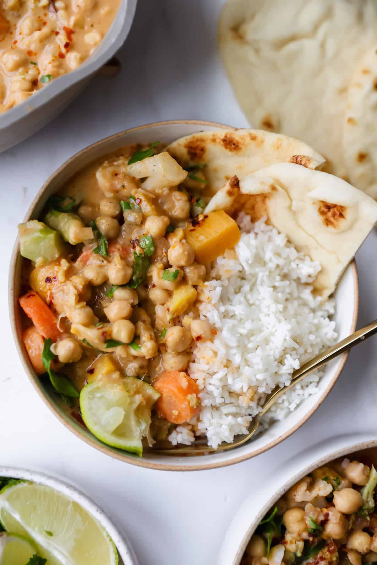 A bowl of chickpea curry with vegetables, white rice, lime wedge, and naan bread, served with a spoon on a white surface.