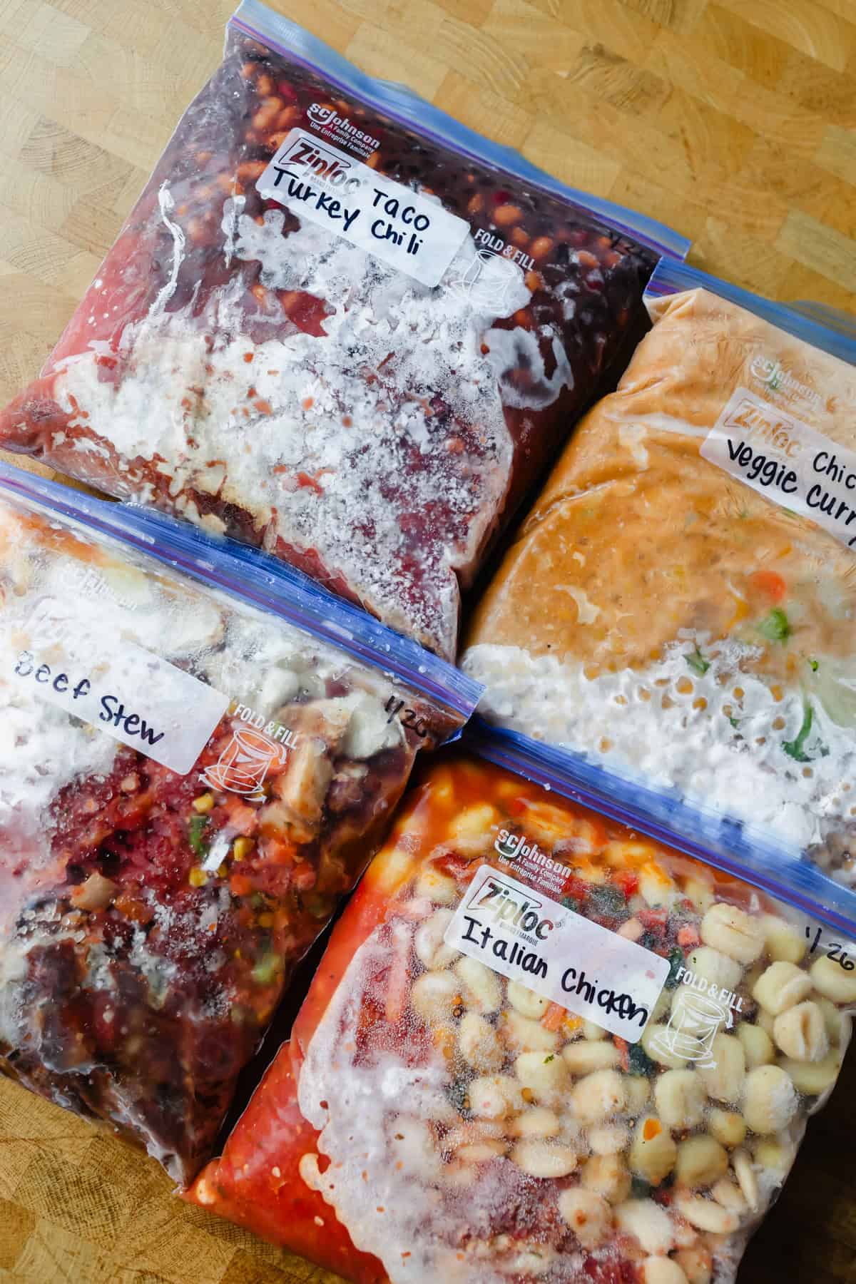 Four labeled freezer bags containing frozen meals: taco turkey chili, veggie curry, beef stew, and Italian chicken, placed on a wooden surface.