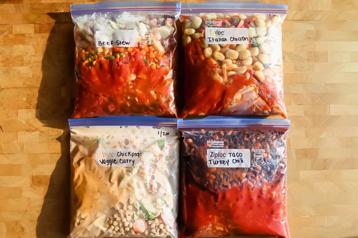 Four labeled freezer bags filled with homemade meals&mdash;beef stew, Italian chicken, chickpea veggie curry, and taco turkey chili&mdash;arranged on a wooden surface.