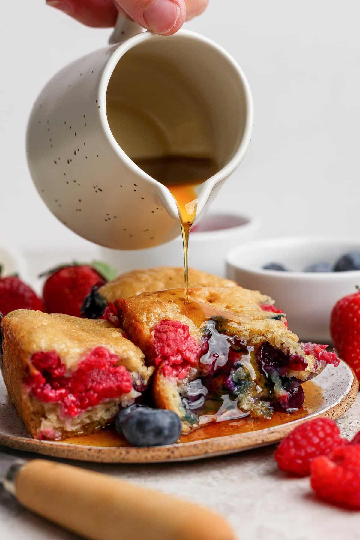 A hand pours syrup from a small white jug onto a stack of berry-filled pancakes on a plate, surrounded by fresh strawberries, raspberries, and blueberries.
