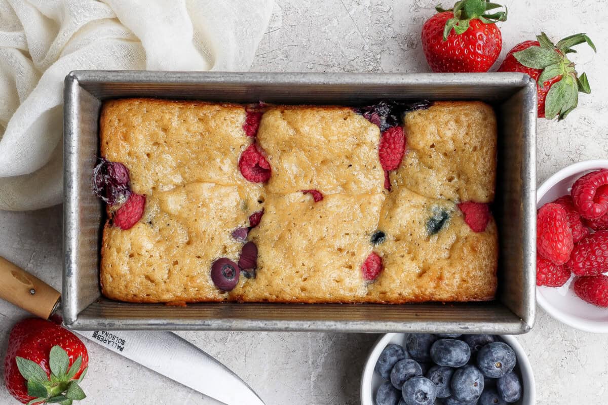 A rectangular loaf pan contains a baked berry bread with visible raspberries and blueberries. Fresh strawberries, blueberries, and raspberries are placed around the pan.