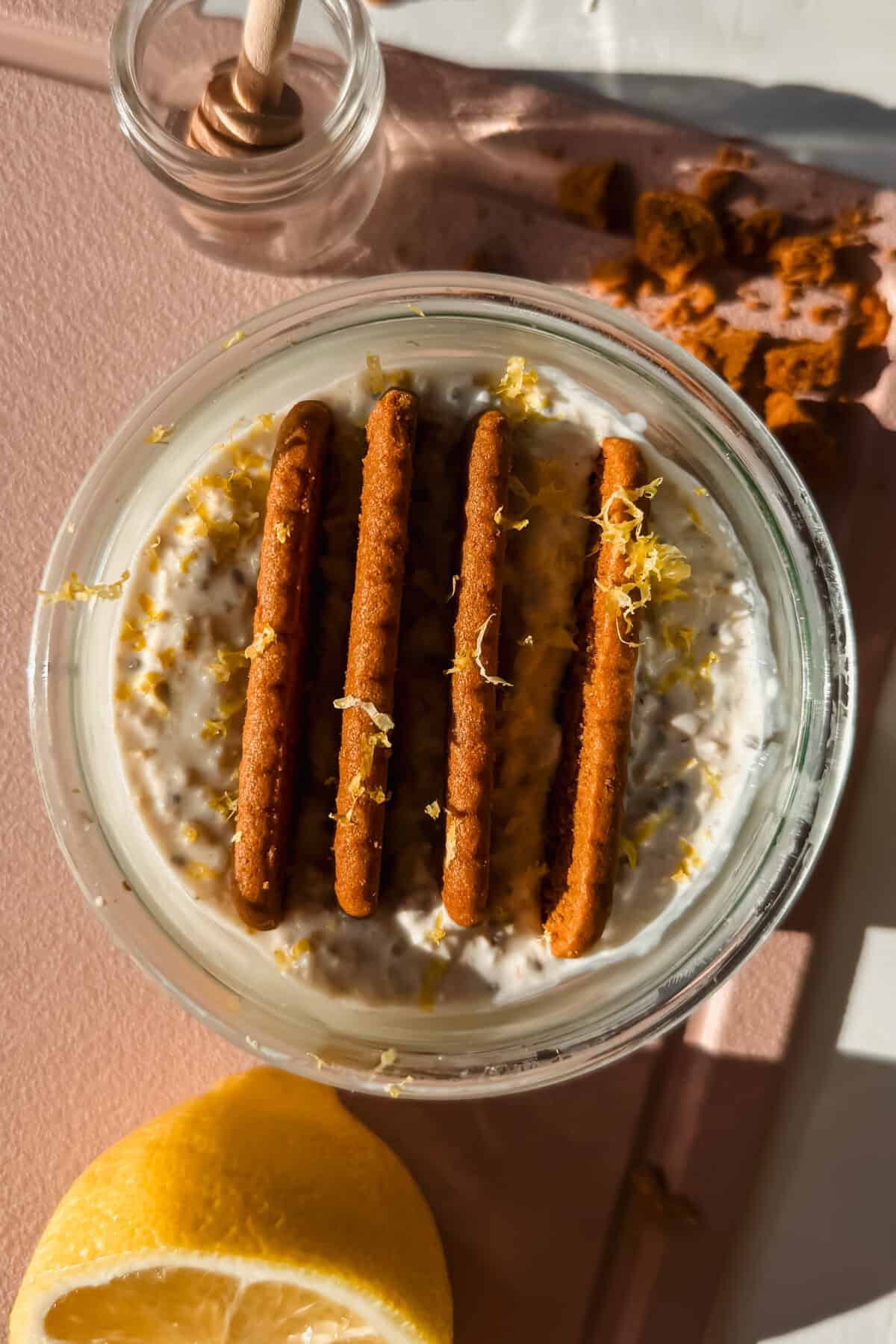 A glass jar of creamy overnight oats is topped with four rectangular cookies and lemon zest, shown with a honey dipper and lemon half nearby on a pink surface.