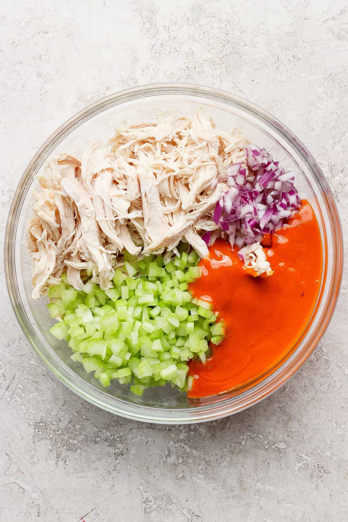A glass bowl containing shredded chicken, chopped celery, diced red onion, and orange buffalo sauce, arranged in separate sections before mixing.