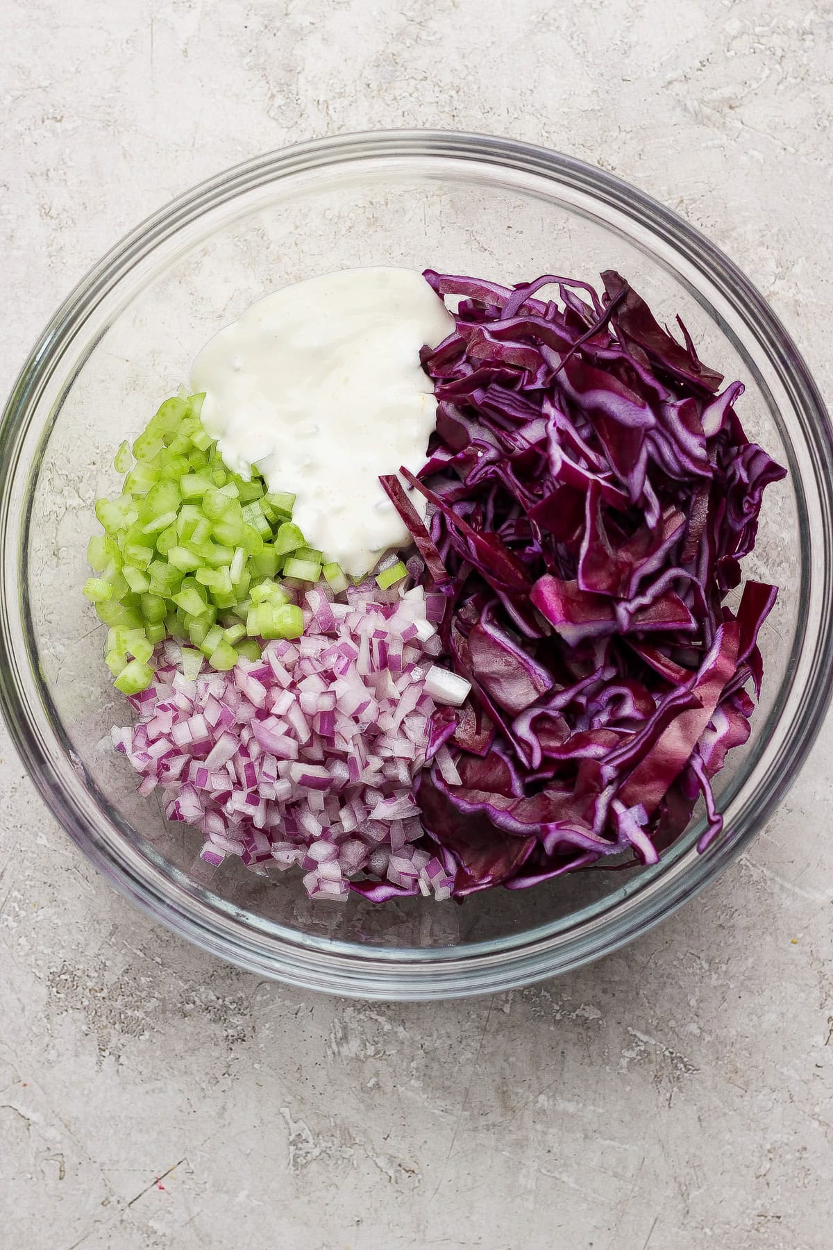 A glass bowl containing chopped celery, chopped red onion, shredded red cabbage, and a dollop of mayonnaise on a light surface.