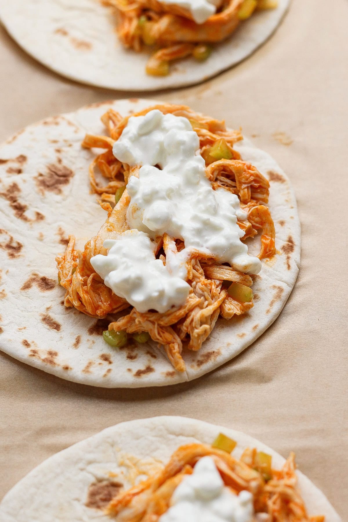 Three flour tortillas topped with shredded chicken, diced vegetables, and a dollop of white sauce, arranged on a light brown surface.