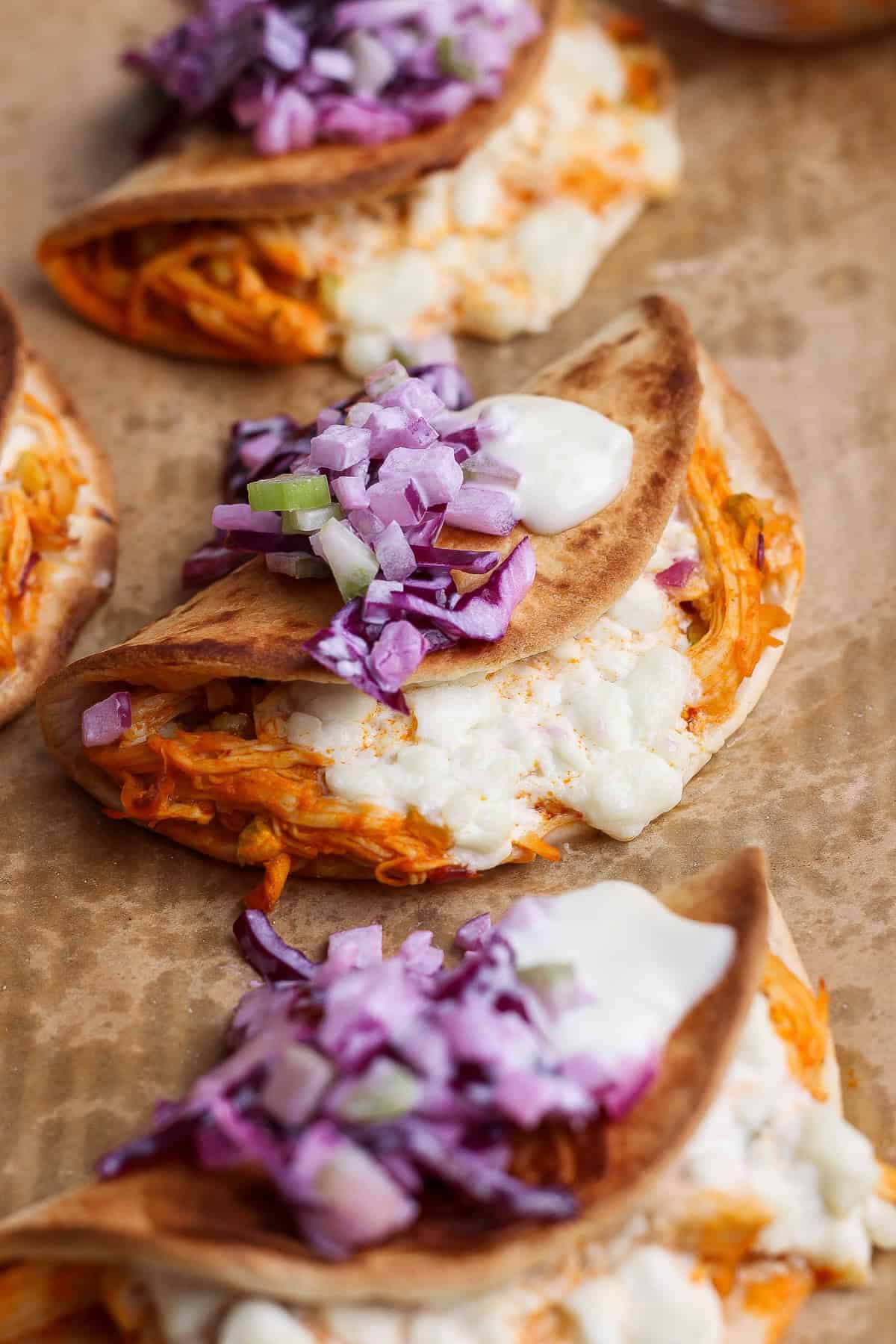 Three folded tacos filled with shredded meat and melted cheese, topped with chopped red onions and a dollop of white sauce, placed on brown parchment paper.