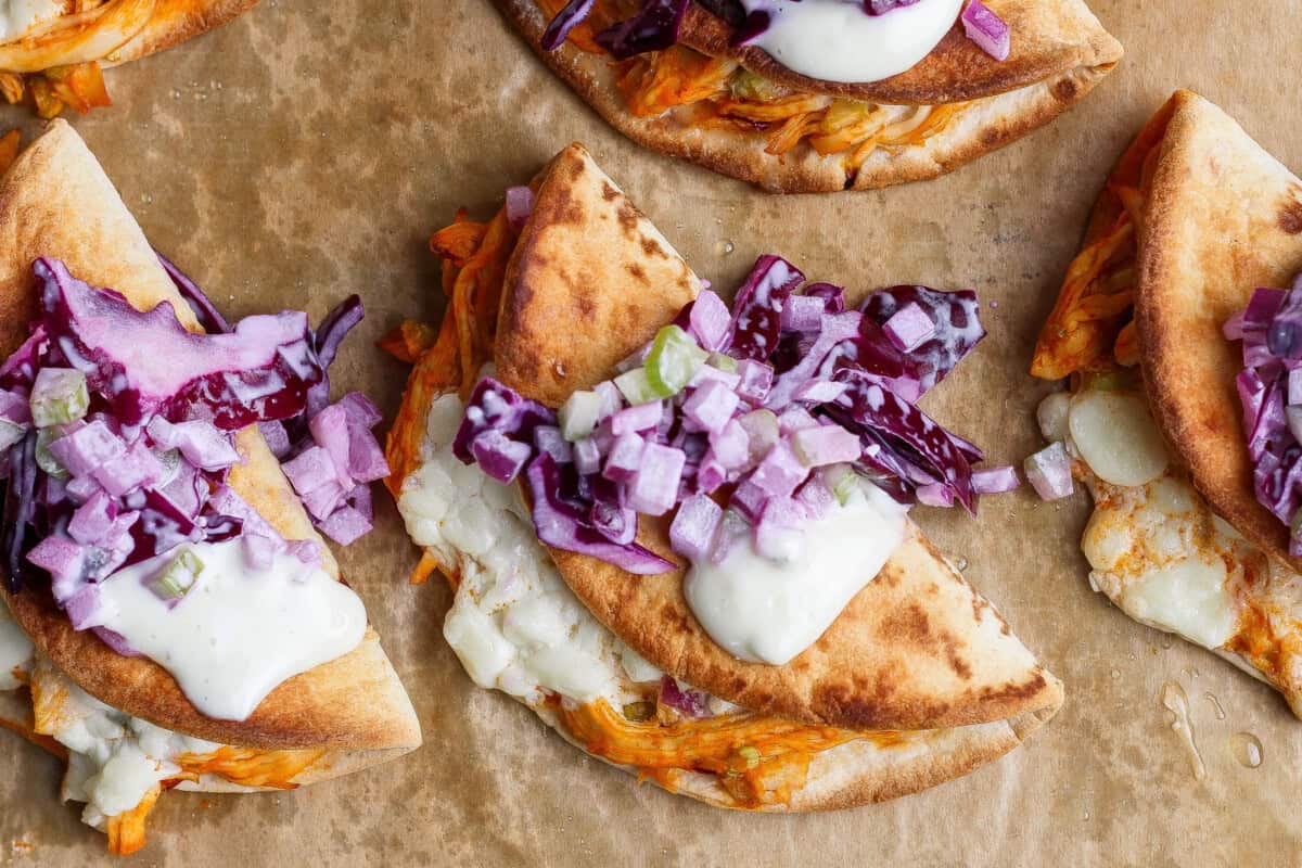 Three folded flatbreads filled with shredded meat, melted cheese, sauce, and topped with chopped red onions and purple cabbage on parchment paper.