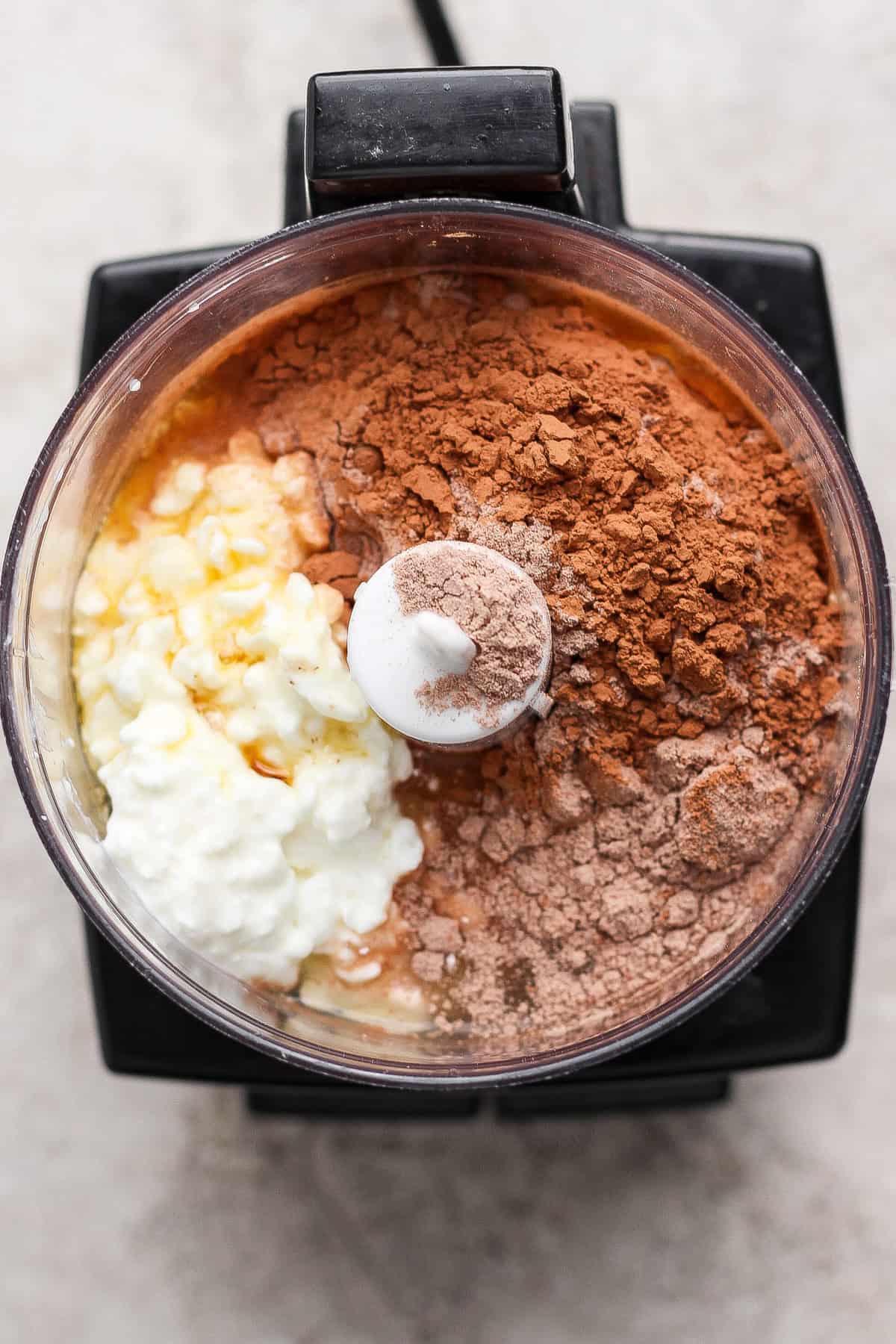 Top view of a food processor bowl containing cottage cheese, cocoa powder, protein powder, and other ingredients before blending.