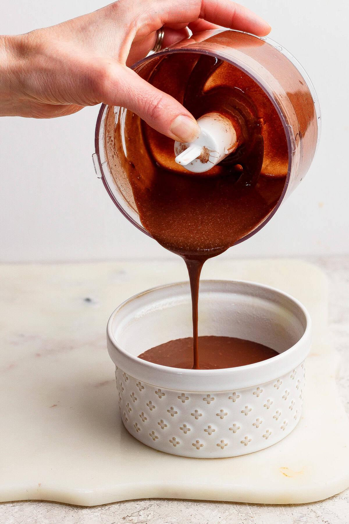 A hand pours thick chocolate sauce from a small food processor bowl into a white ceramic ramekin on a light surface.