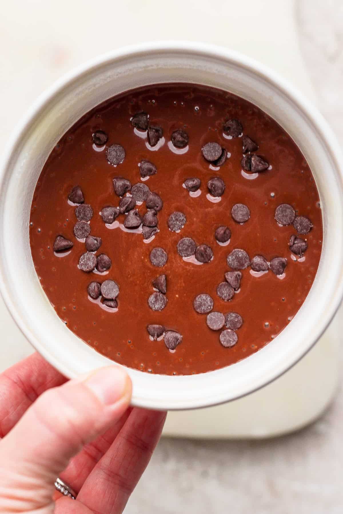 A hand holds a white bowl filled with chocolate batter topped with scattered chocolate chips.