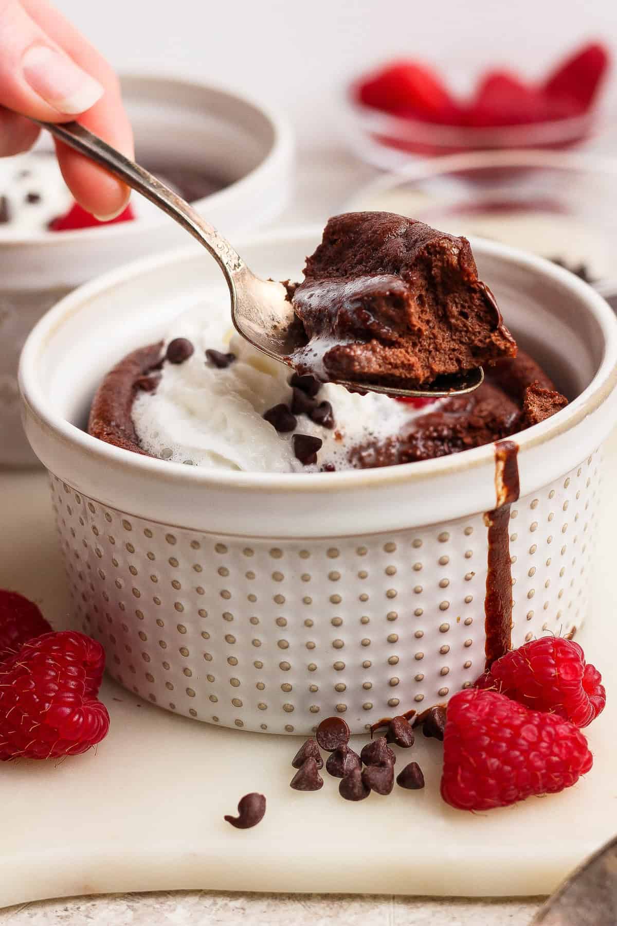 A hand holds a spoonful of chocolate lava cake topped with whipped cream in a white ramekin, with raspberries and chocolate chips on the side.