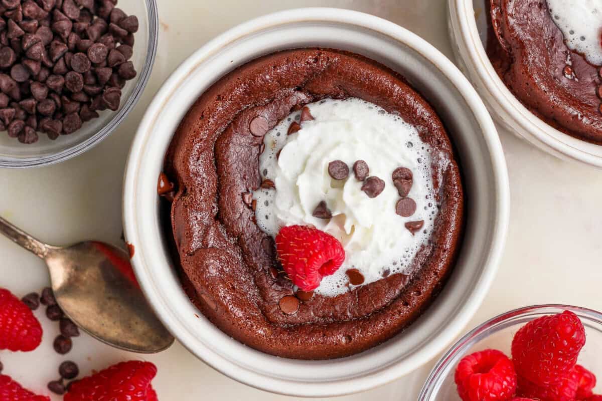 A chocolate lava cake in a white ramekin topped with whipped cream, chocolate chips, and a raspberry, surrounded by bowls of chocolate chips and raspberries.