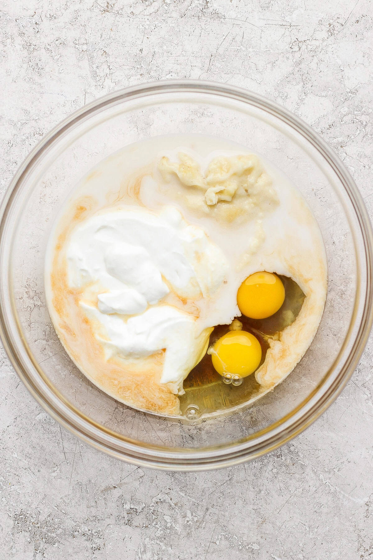 A glass bowl containing two raw eggs, yogurt, and other wet ingredients on a light textured surface.