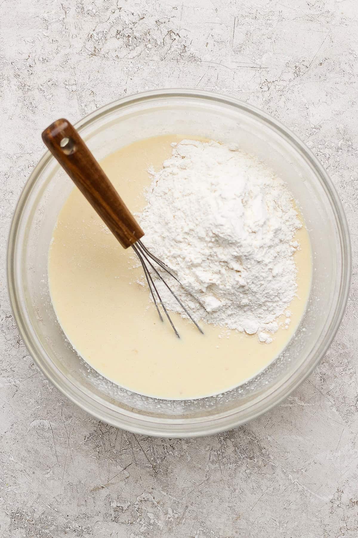 A glass bowl with liquid batter and a mound of flour, being mixed with a metal whisk with a wooden handle, on a light textured surface.