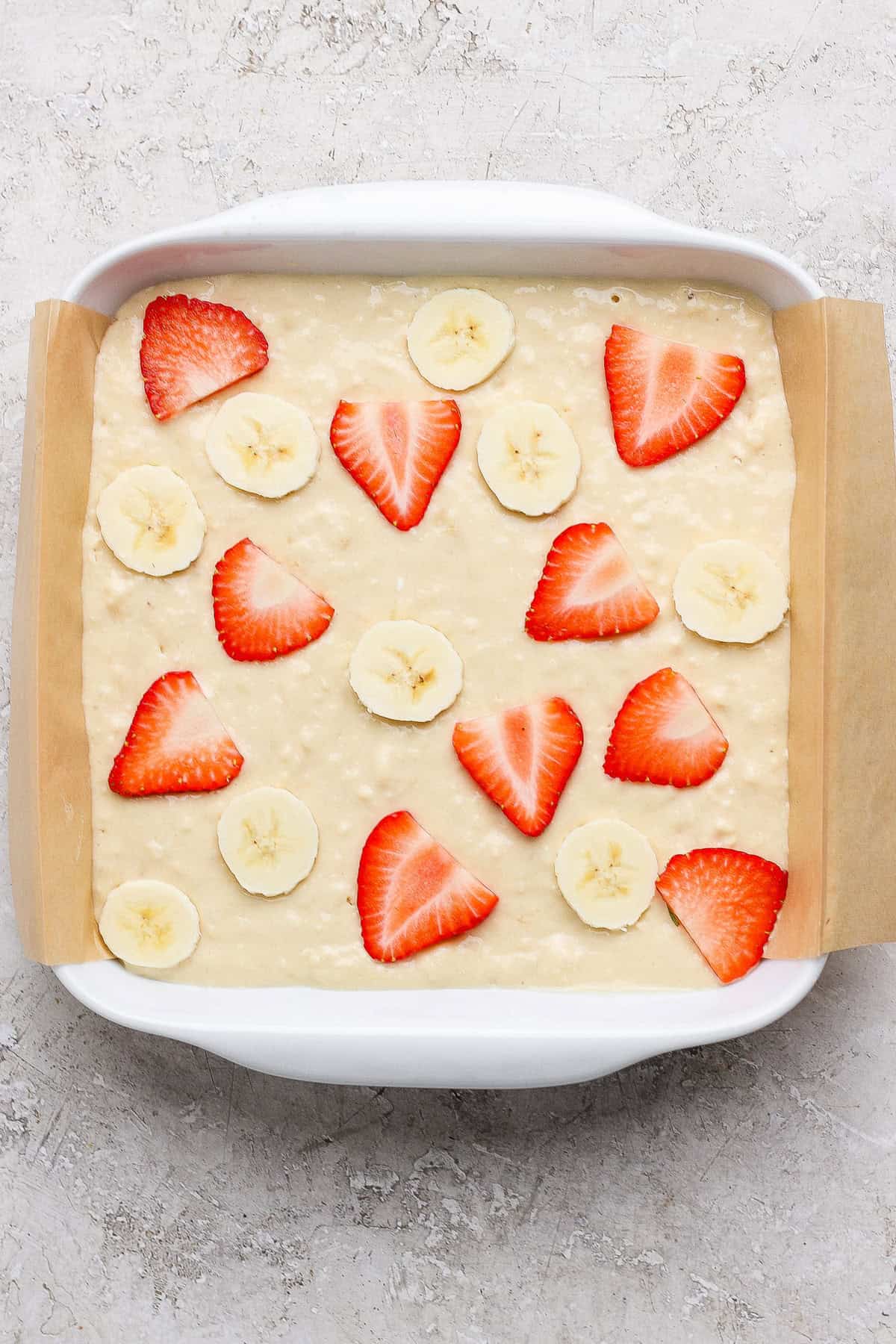 A baking dish lined with parchment paper contains unbaked batter topped with sliced strawberries and bananas.