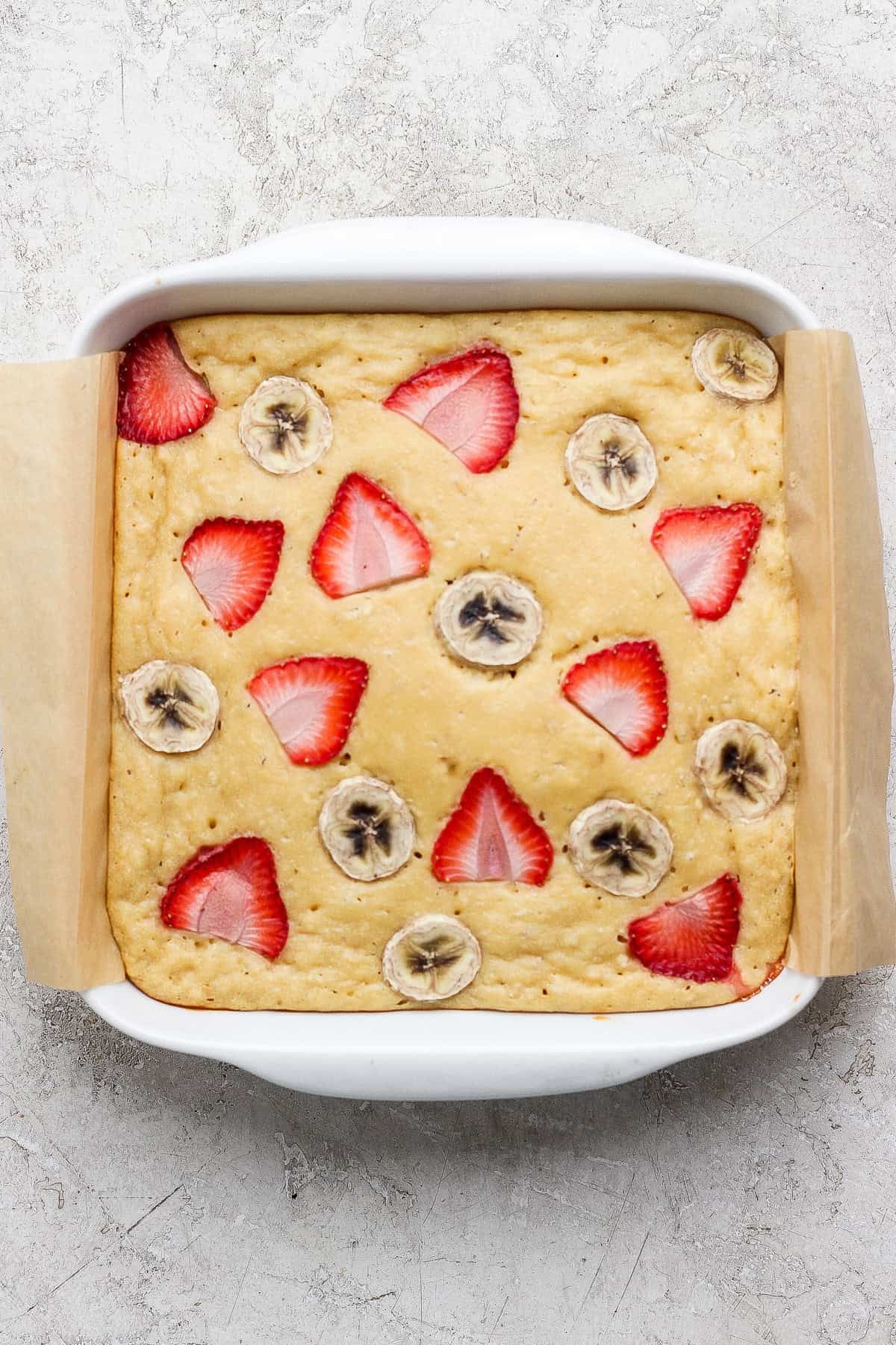 A square baking dish with a baked cake topped with sliced strawberries and bananas, lined with parchment paper on a light textured surface.