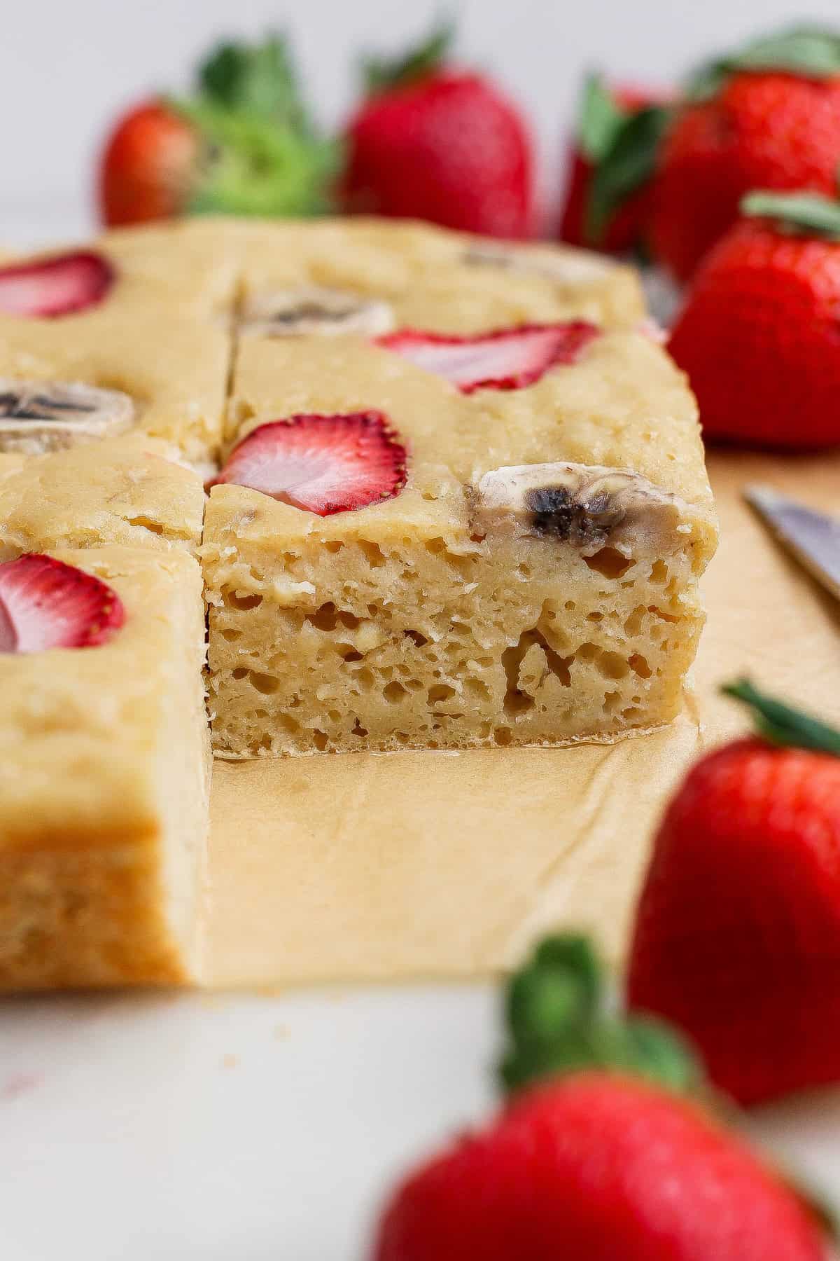 A sponge cake topped with sliced strawberries and bananas sits on parchment paper, with a slice cut out and whole strawberries around it.