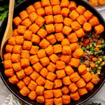 A skillet filled with a layer of sweet potato tater tots on top of a ground meat and mixed vegetable filling, with a wooden spoon resting on the side.