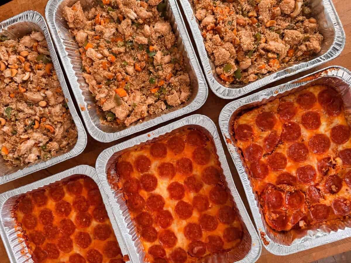 Six aluminum trays, three filled with pepperoni pizza and three with a stir fry dish of vegetables and chicken, are arranged on a wooden surface&mdash;perfect alongside comforting casseroles for any gathering.