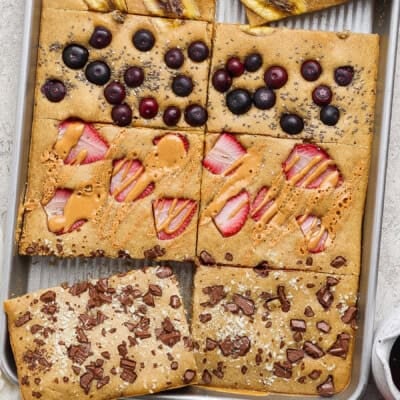 A sheet pan pancake divided into sections, each topped with different ingredients: banana, blueberries, peanut butter with strawberries, and chocolate pieces.