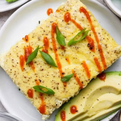 A rectangular egg dish garnished with sliced green onions and hot sauce, served with avocado slices on a white plate.