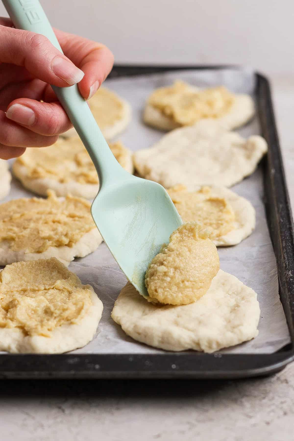 A hand uses a pale green spatula to spread cottage cheese filling onto rounds of dough for pull apart bread, all arranged on a baking sheet lined with parchment paper.