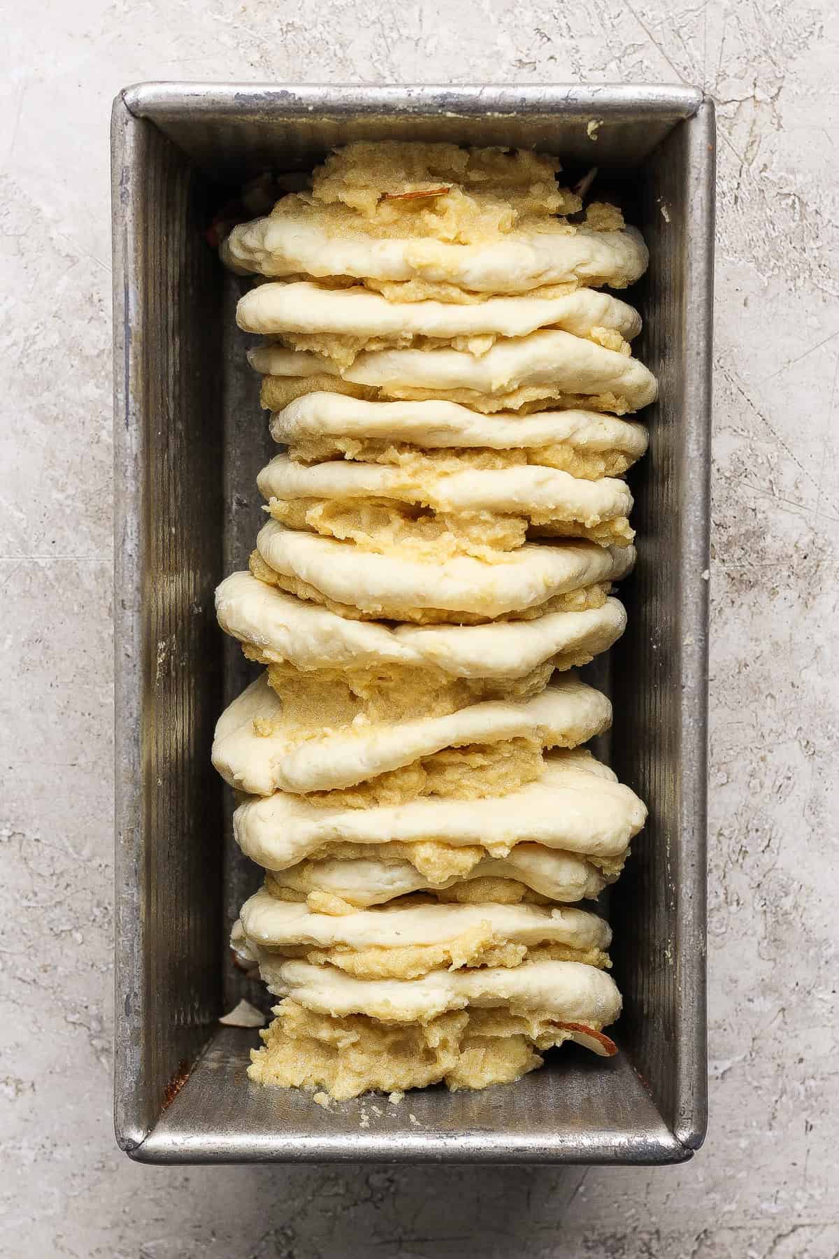 A metal loaf pan with stacked layers of unbaked dough and a yellow cottage cheese filling, arranged vertically for delicious pull apart bread, ready to bake.