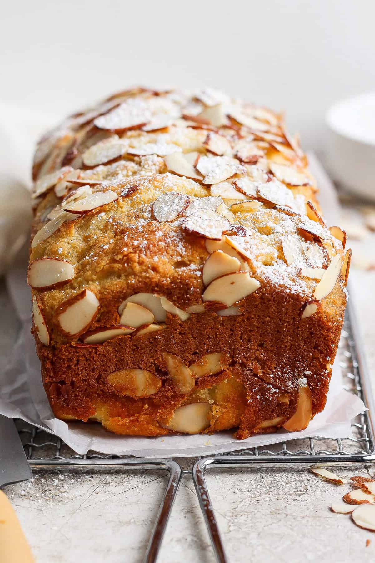 A loaf of almond cake topped with sliced almonds and powdered sugar sits on parchment paper next to a cottage cheese pull apart bread on a cooling rack.