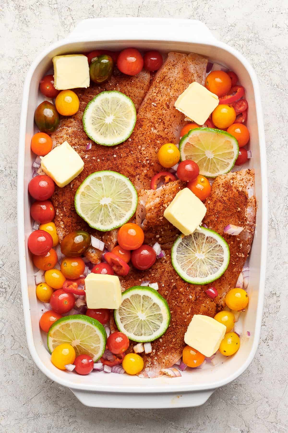 Raw seasoned fish fillets in a baking dish topped with lime slices, cherry tomatoes, onions, and butter cubes, ready for cooking.