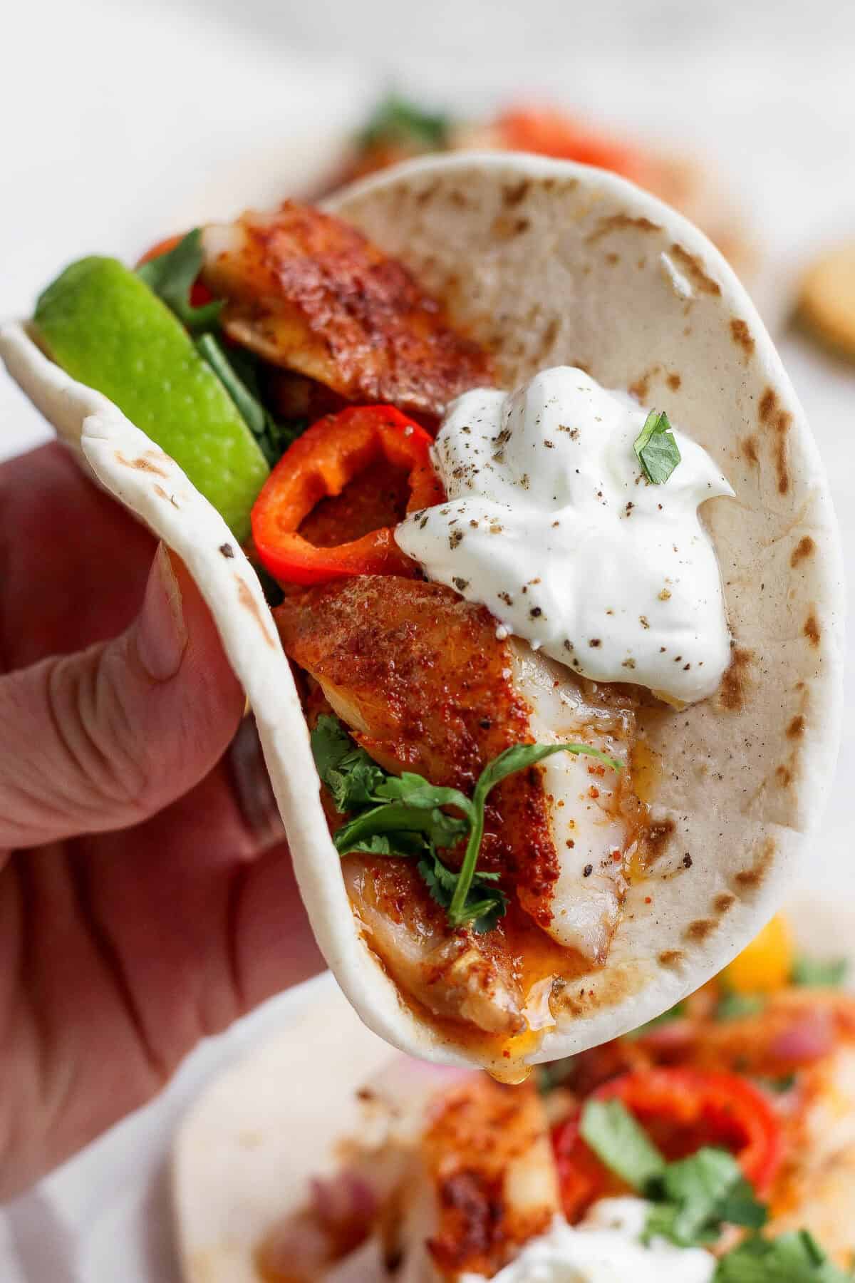 A hand holds a soft taco filled with seasoned fish, sliced red peppers, fresh greens, avocado, and a dollop of sour cream, garnished with black pepper and herbs.