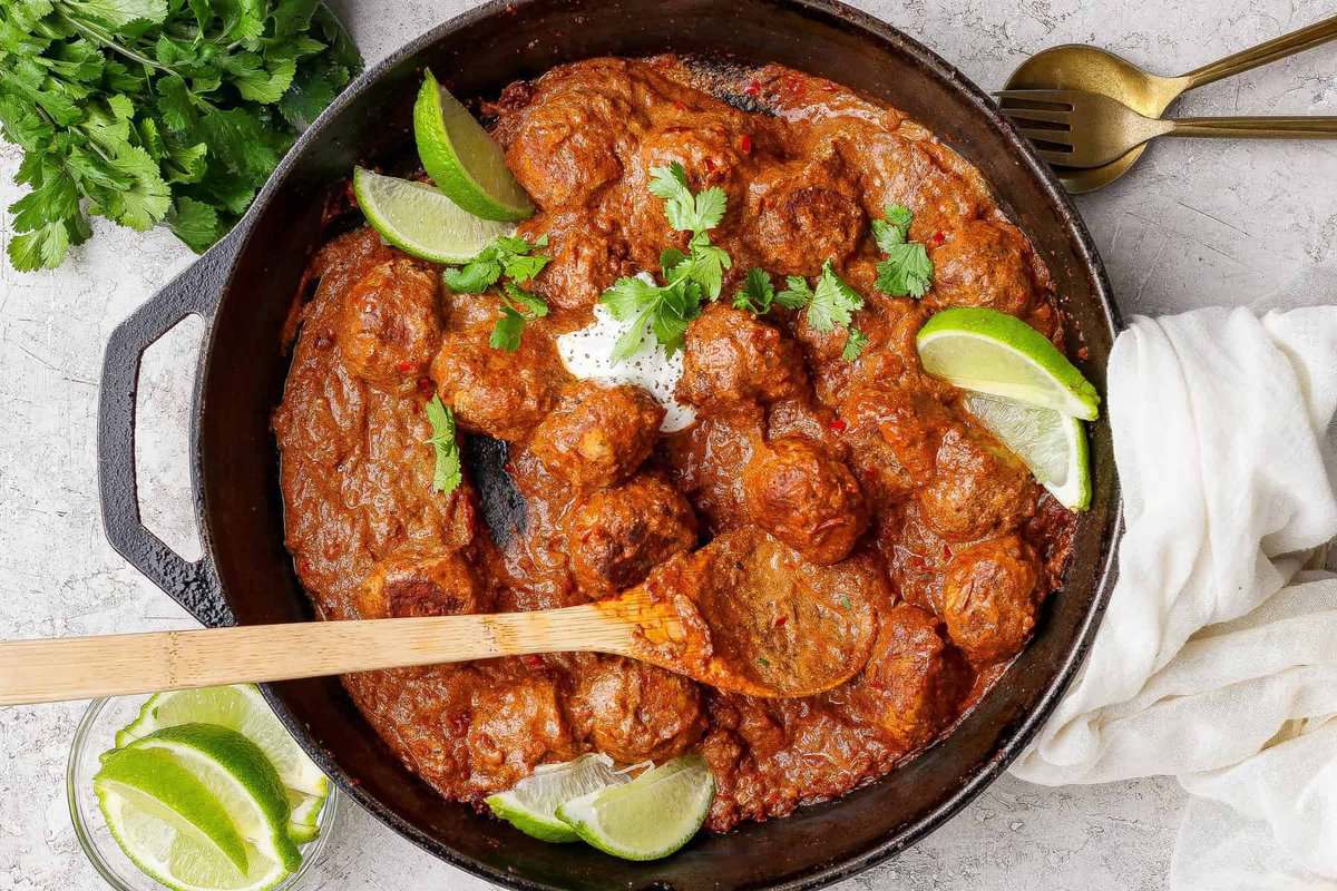 A cast iron skillet filled with butter chicken meatballs in a rich red sauce, garnished with lime wedges and cilantro, with a wooden spoon resting inside. Fresh cilantro and lime are on the side.