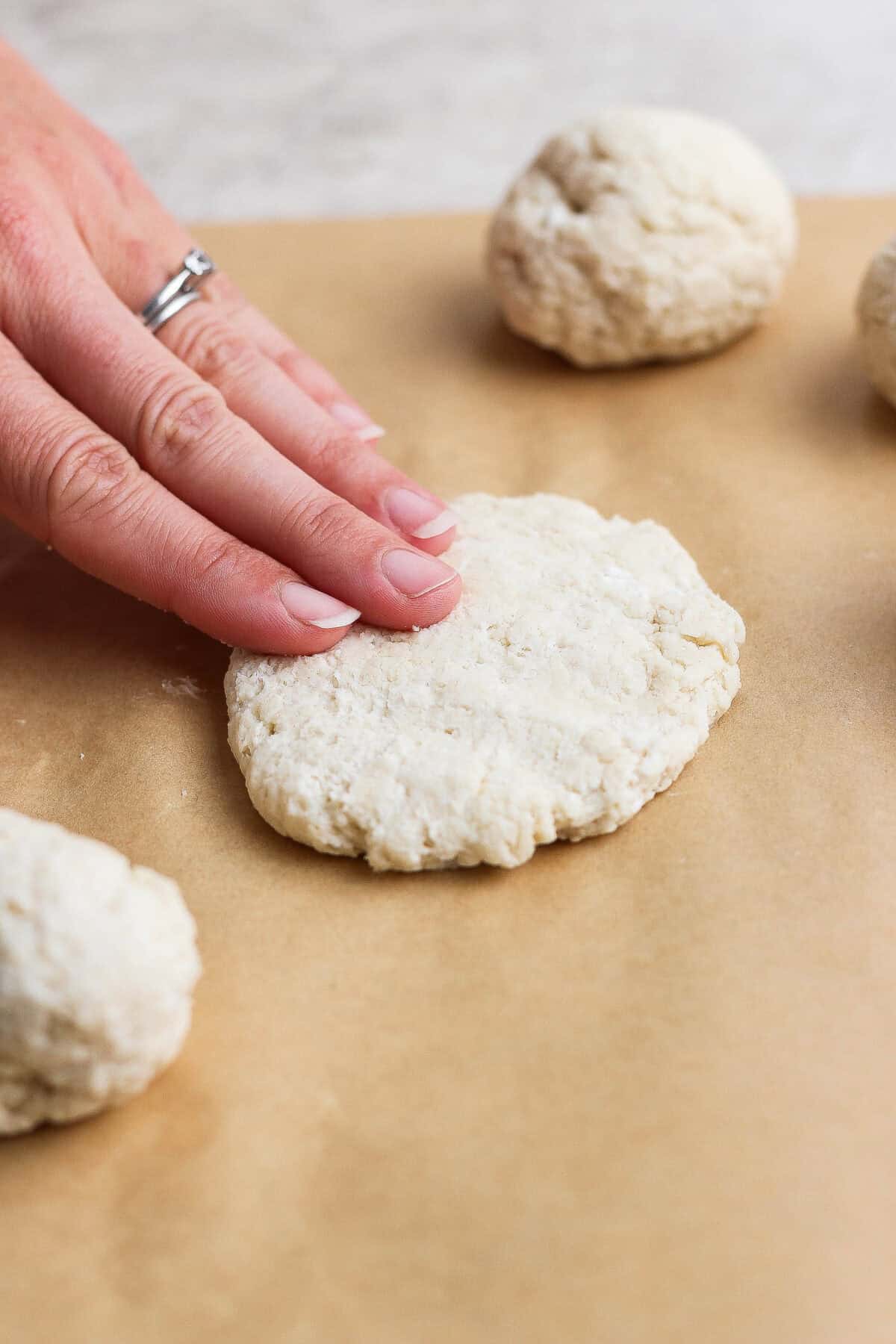 A hand presses a round piece of dough flat on parchment paper, shaping garlic naan, with other dough balls nearby, preparing biscuits.