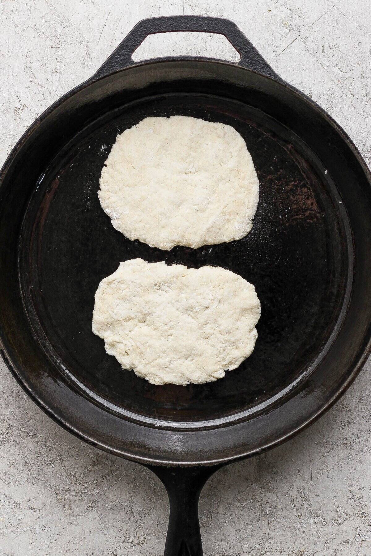 Two pieces of raw garlic naan dough are placed side by side in a large black cast iron skillet on a light textured surface.