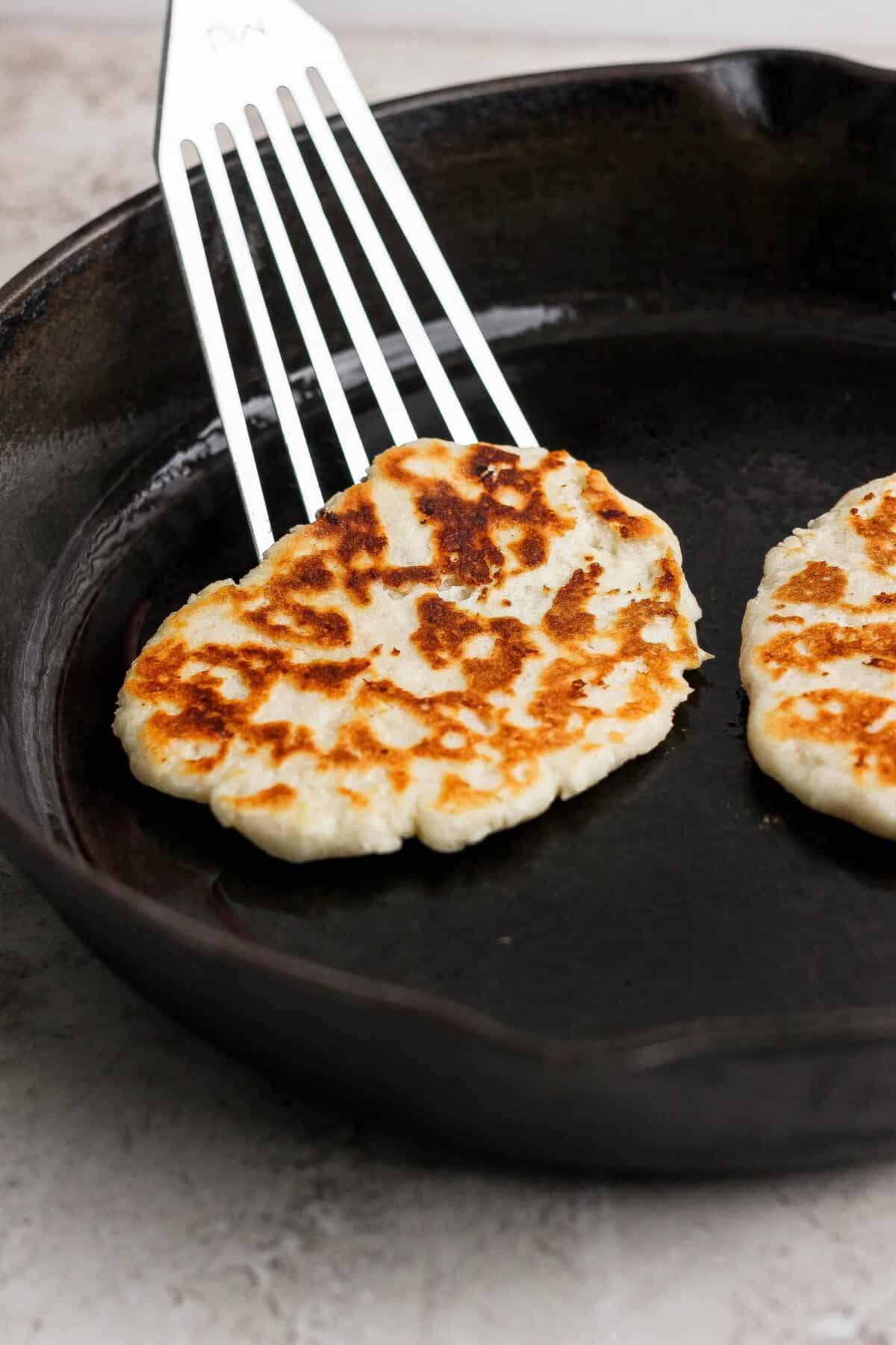 A metal spatula lifts a browned garlic naan flatbread from a cast iron skillet, with another naan still in the pan.