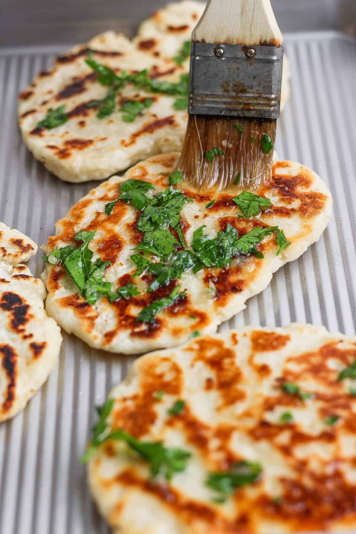 A basting brush is spreading oil and chopped herbs onto freshly cooked garlic naan on a baking sheet.