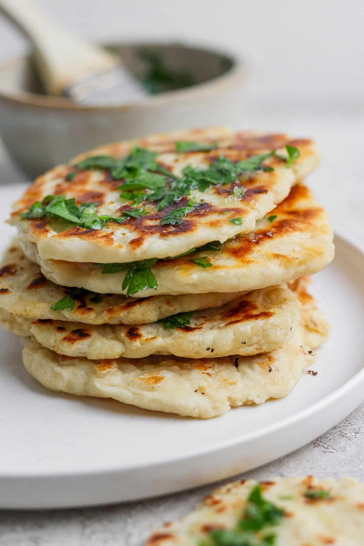 A stack of golden-brown garlic naan flatbreads garnished with chopped herbs, served on a white plate.