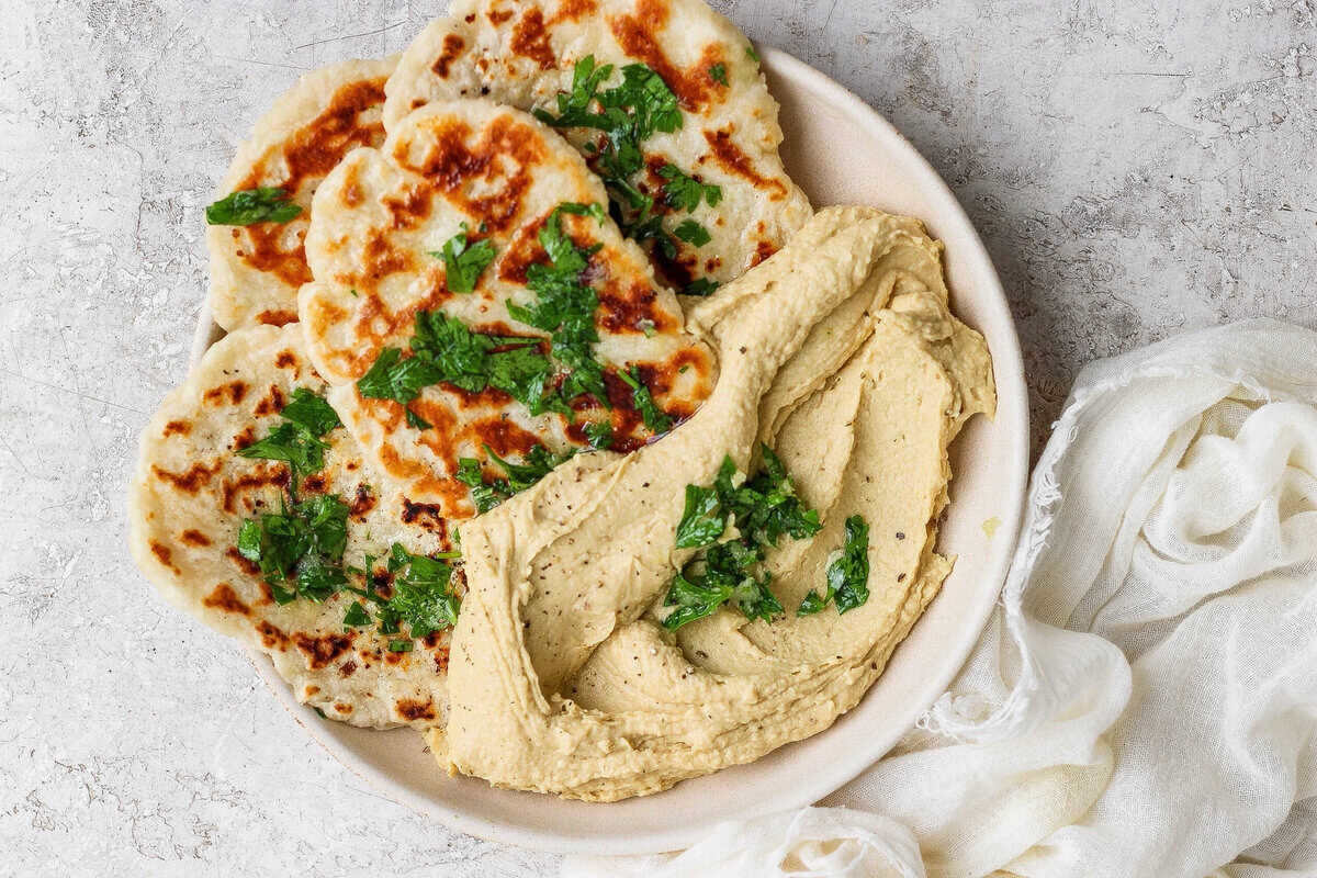 A plate with grilled garlic naan flatbreads topped with chopped herbs, served beside a generous portion of hummus, on a light textured background.
