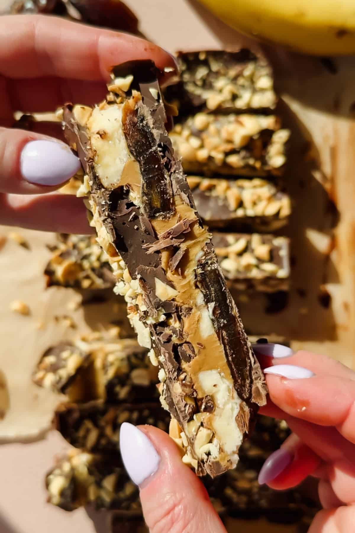 A person holding a chocolate and nut bar filled with banana, peanut butter, and nuts; sliced bars are arranged on parchment paper in the background.
